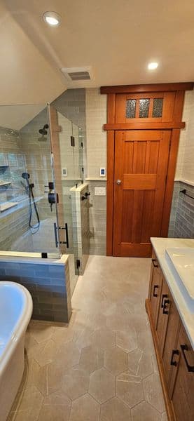 Modern bathroom with glass shower, wooden door, hexagonal tiles, and dual sinks.
