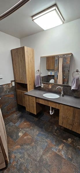 Modern bathroom with wooden cabinetry, stone floor, and large mirror reflecting bright lighting.