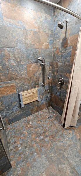 Accessible shower with textured stone walls, handheld showerhead, and foldable bench.